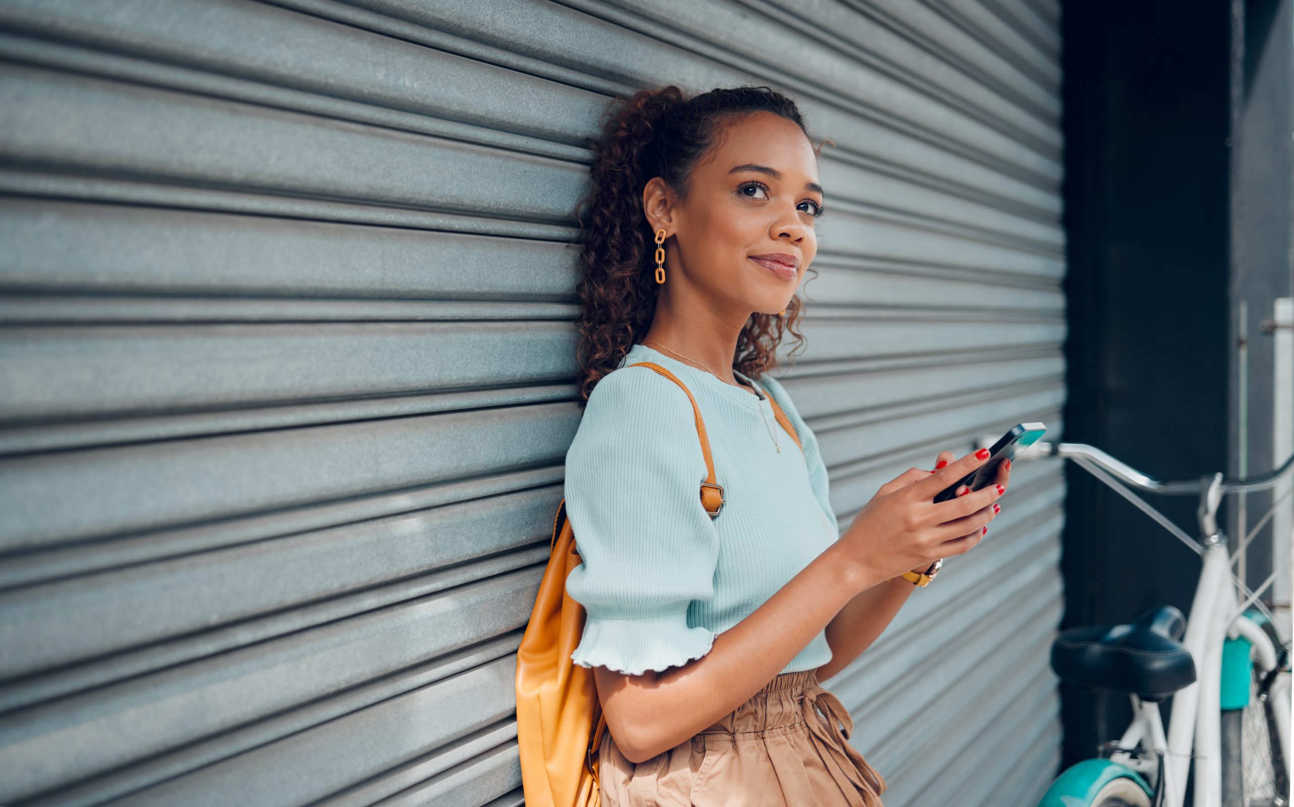 City lifestyle, girl with phone and bike standing at wall thinking before writing text. Happy female student, bicycle and smartphone outside street, communication, 5g and sustainable urban transport.